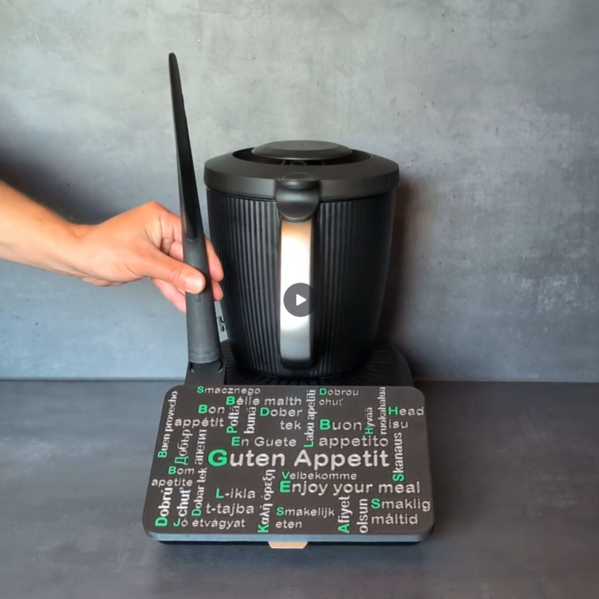 TM7 Displayschutz mit mehrsprachigem ‚Guten Appetit‘-Schriftzug. Gezeigt wird das Aufsetzen der Displayabdeckung auf den Thermomix TM7 sowie das Einsetzen des Spatels in den integrierten Spatelhalter.